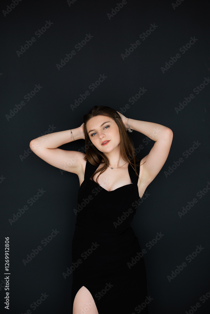 woman with long light brown hair. She is wearing a black off-the-shoulder dress with a plunging neckline. Woman standing against a dark wall woman with long light brown hair. She is wearing a black off-the-shoulder dress with a plunging neckline. Woman standing against a dark wall