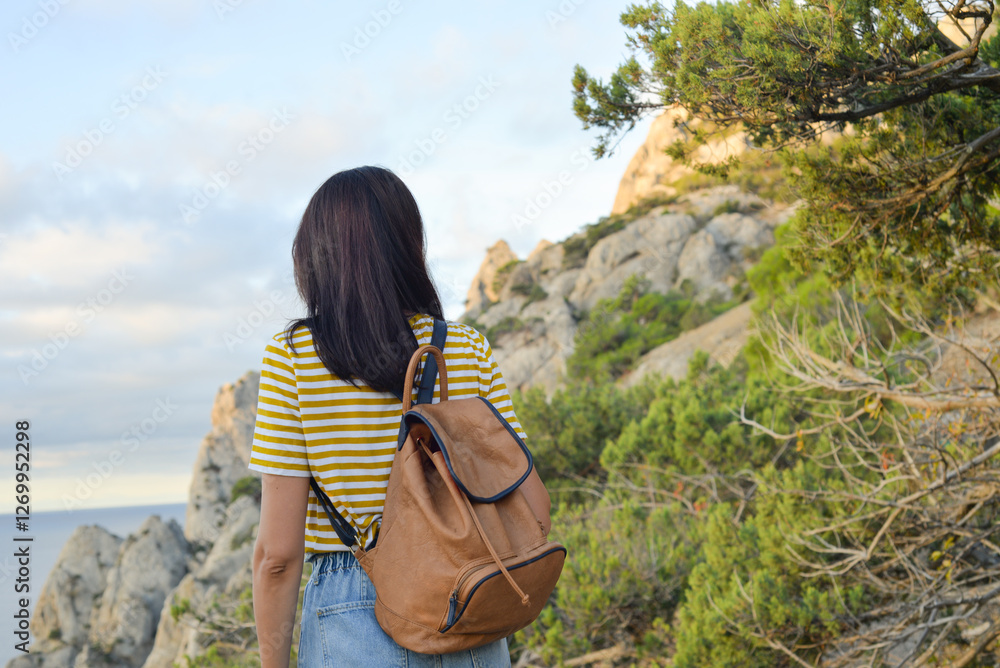 Happy female tourist with a backpack on the top of a mountain, enjoying the sea view, standing on a rock and looking into the distance. The village of Novy Svet in Crimea. Russia