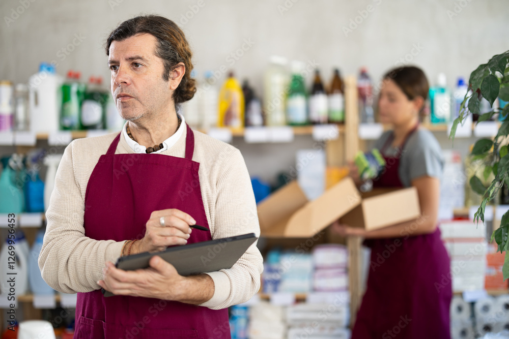 Experienced household goods store owner using tablet to manage inventory of cleaning supplies and toiletries, while female worker organizing products on shelves in background Experienced household goods store owner using tablet to manage inventory of cleaning supplies and toiletries, while female worker organizing products on shelves in background