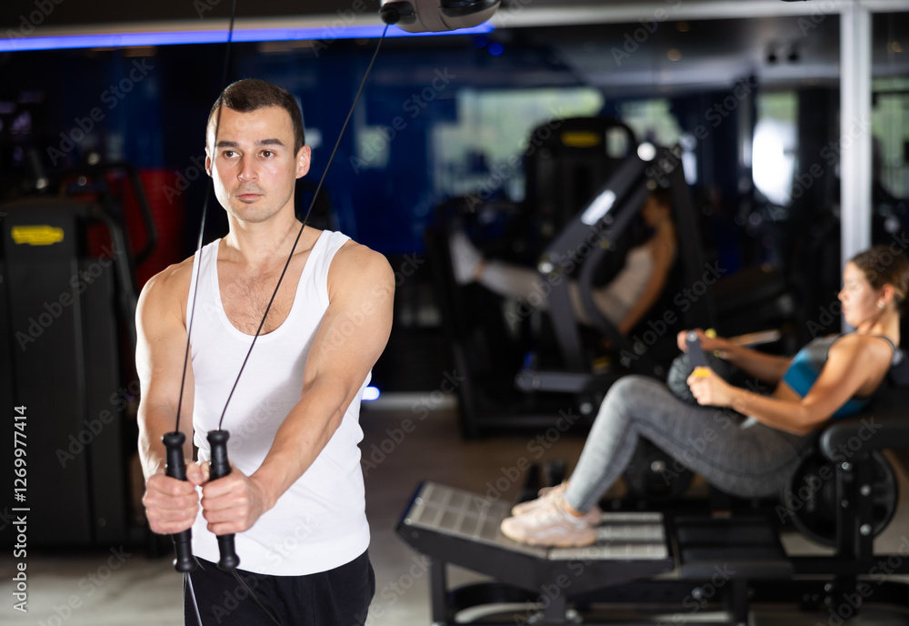 Fototapeta premium Slender athletic man work out muscles of arms and shoulders on TRX rope loop simulator in gym of fitness club. Gym visitor trains triceps during classes at multi-station in wellness center.
