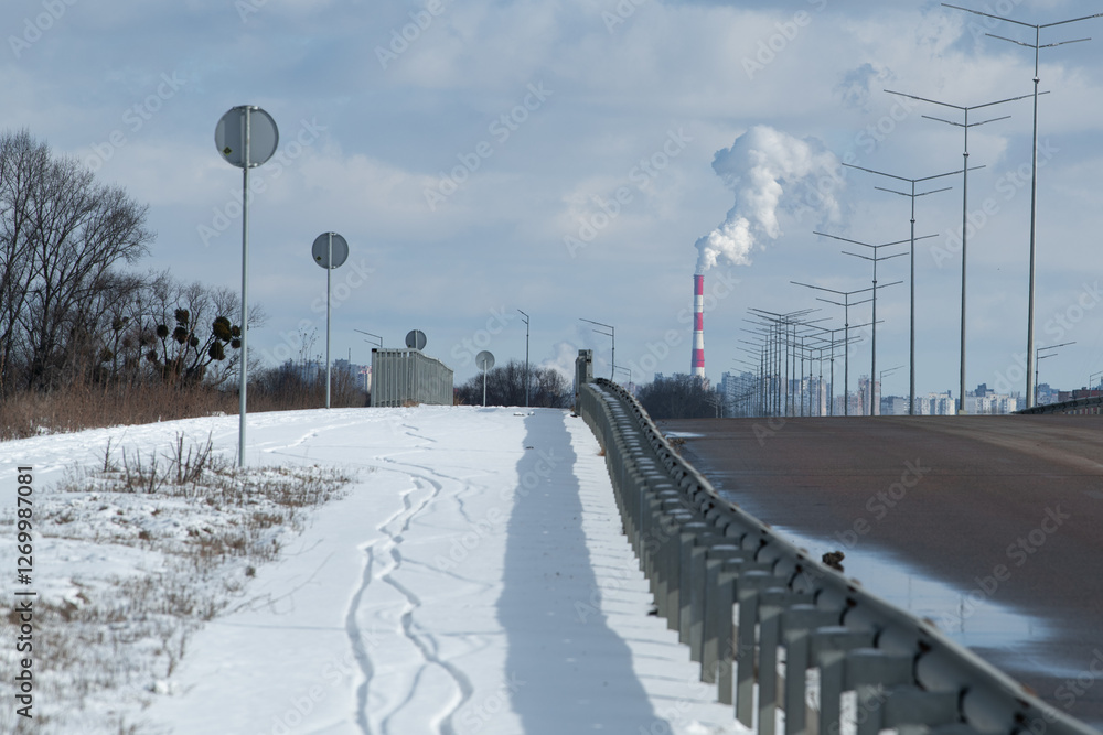 A snow-covered roadside showcases a winter landscape, with a barrier separating the snowy path from a wide road. Industrial smokestacks release clouds of smoke against a gray sky A snow-covered roadside showcases a winter landscape, with a barrier separating the snowy path from a wide road. Industrial smokestacks release clouds of smoke against a gray sky