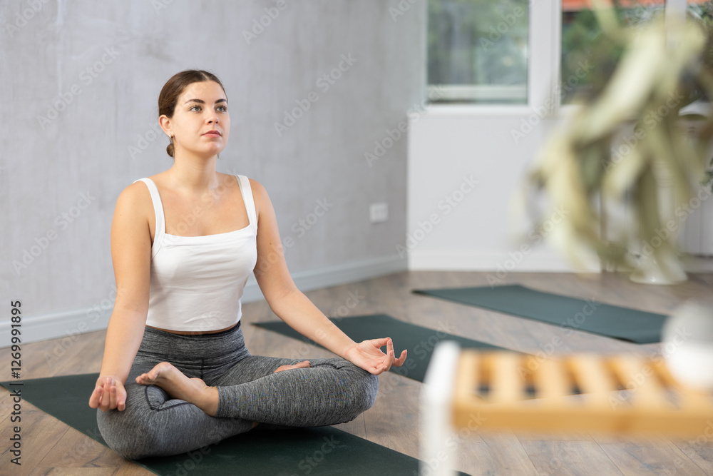 Positive young girl meditating in modern yoga studio, sitting in lotus position with hands on knees with fingers folded in mudra Positive young girl meditating in modern yoga studio, sitting in lotus position with hands on knees with fingers folded in mudra