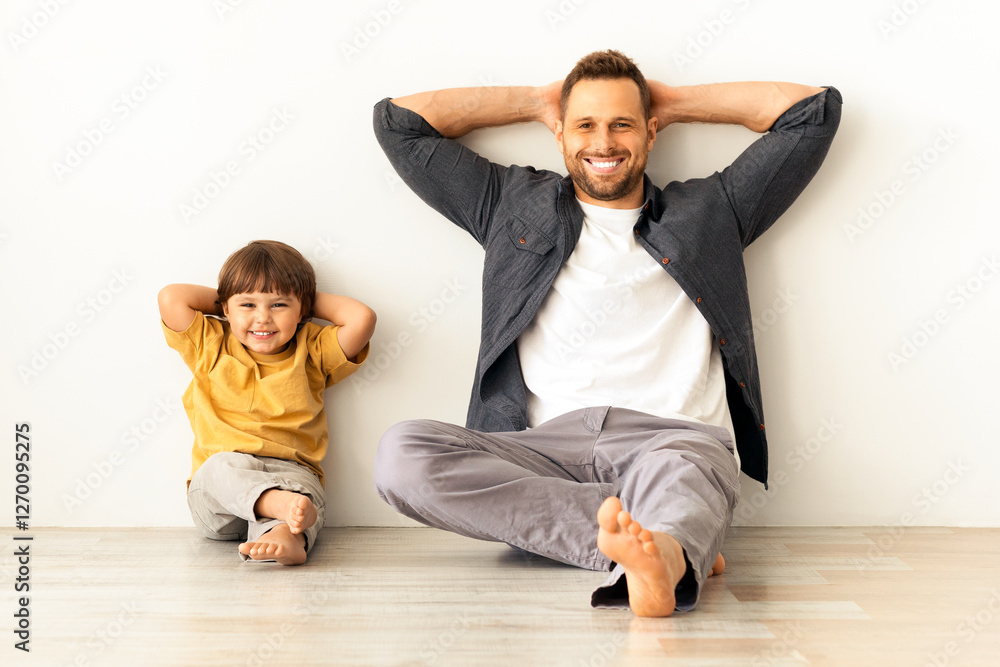 Friendly family. Happy relaxed father and his little adorable son sitting on floor near wall and smiling to camera, expressing peace and relax Friendly family. Happy relaxed father and his little adorable son sitting on floor near wall and smiling to camera, expressing peace and relax