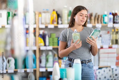 Young female customer in home essentials store holding two bottles of dishwashing liquid, carefully comparing product features before making decision photo