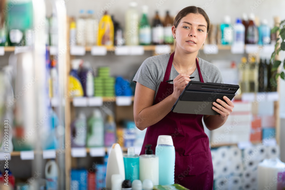 Smiling confident young female household goods store worker using tablet to manage inventory of cleaning supplies and toiletries.. Smiling confident young female household goods store worker using tablet to manage inventory of cleaning supplies and toiletries..