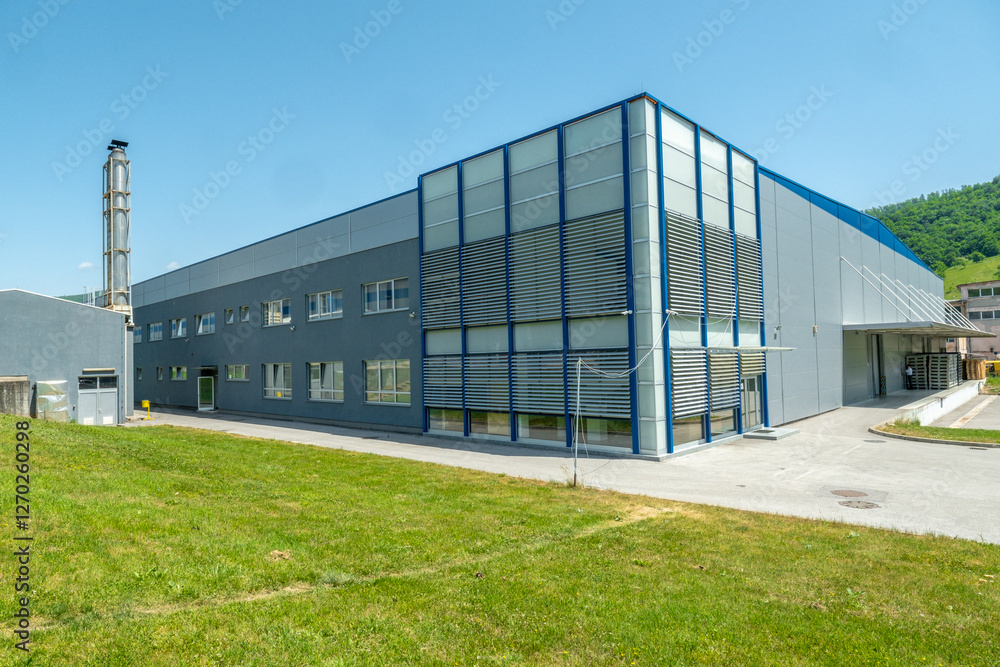 Fototapeta premium Large industrial building with gray and blue exterior under a bright blue sky surrounded by green grass