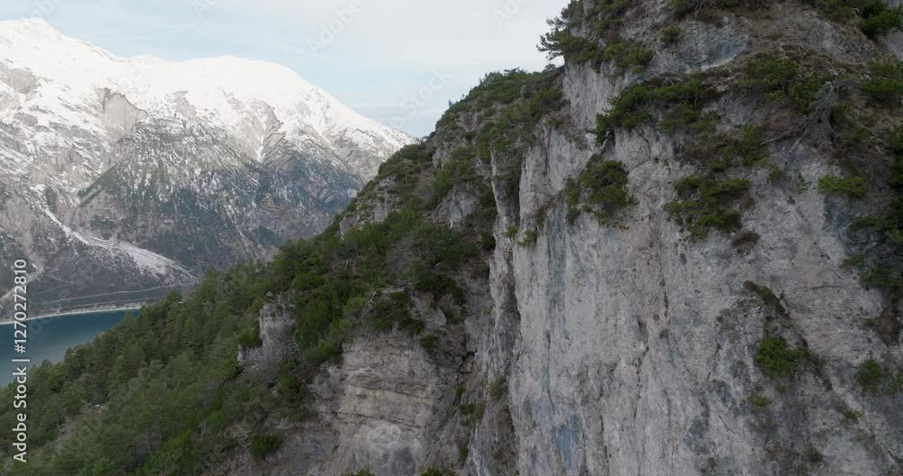 A stunning aerial view of a rocky cliff with sparse greenery, overlooking Achensee Lake. Snow-capped mountains in the background create a dramatic alpine landscape. A stunning aerial view of a rocky cliff with sparse greenery, overlooking Achensee Lake. Snow-capped mountains in the background create a dramatic alpine landscape.