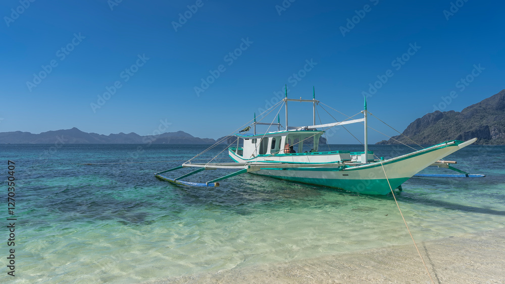 Fototapeta premium A beautiful traditional double-outrigger dugout Filipino bangka boat is moored at the shore. The ropes are stretched. The turquoise ocean water is crystal clear. Mountains on the horizon. Blue sky.