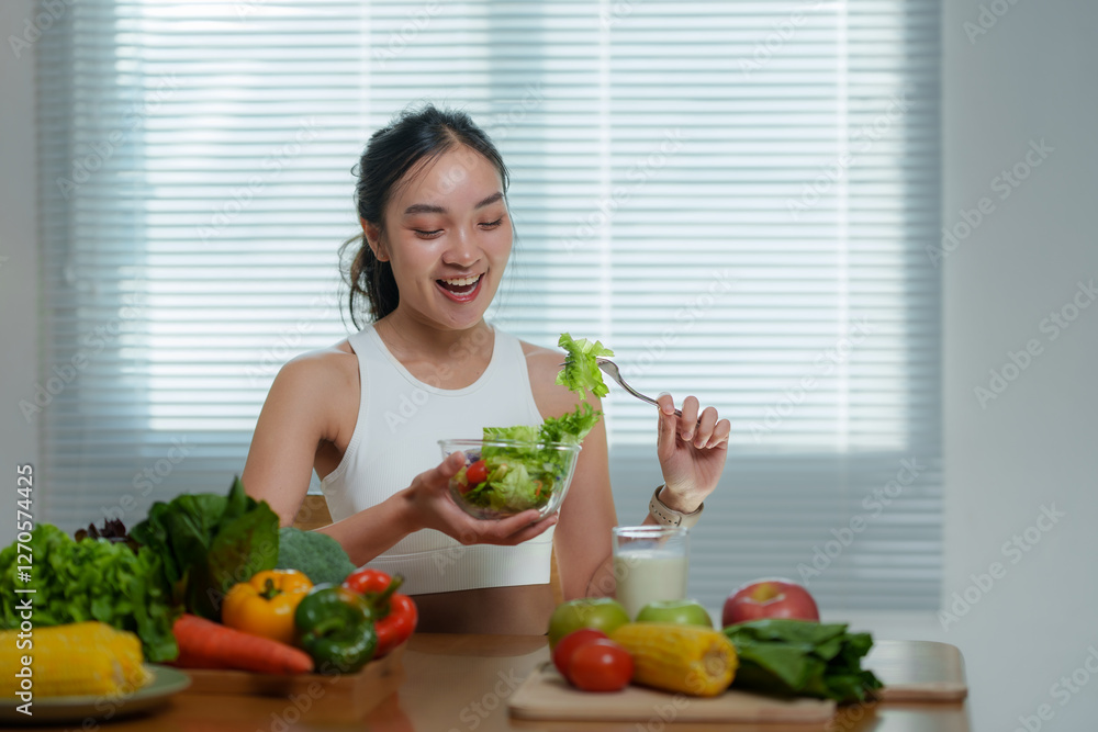 Young smiling sportswoman is eating a bowl of fresh salad sitting at the table at home with a variety of fresh vegetables and fruits on the table, promoting a healthy lifestyle Young smiling sportswoman is eating a bowl of fresh salad sitting at the table at home with a variety of fresh vegetables and fruits on the table, promoting a healthy lifestyle