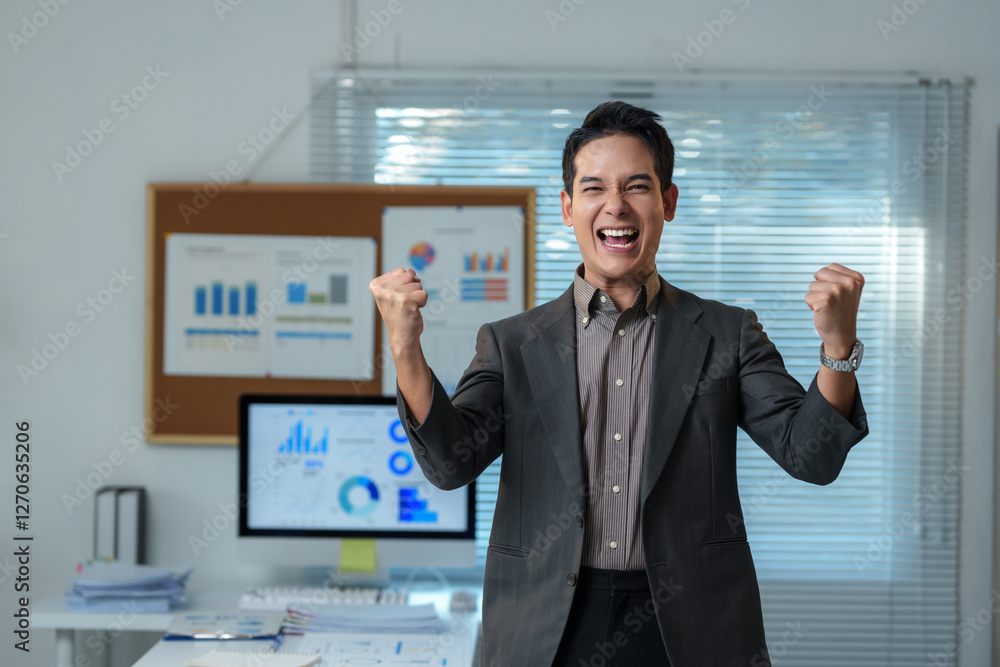 Young businessman is raising his arms and celebrating success in office with charts and graphs on the background, expressing his excitement and achievement Young businessman is raising his arms and celebrating success in office with charts and graphs on the background, expressing his excitement and achievement