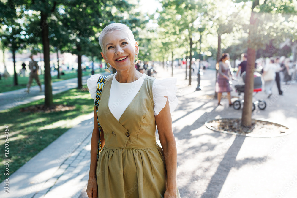 Fototapeta premium Outdoor portrait of stylish grandma walking in park going to meet with her family and grandchildren on playground, looking at camera with smile and joy, wearing trendy dress and imitation jewelry