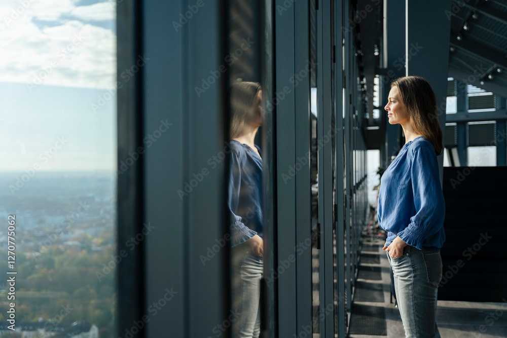 Smiling businesswoman standing with hands in pockets and looking through window at office Smiling businesswoman standing with hands in pockets and looking through window at office