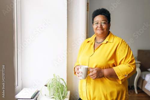 Foto Portrait of overweight elderly female of african ethnicity drinking healthy vitamin tea being on diet standing near window in bedroom, looking at camera with smile