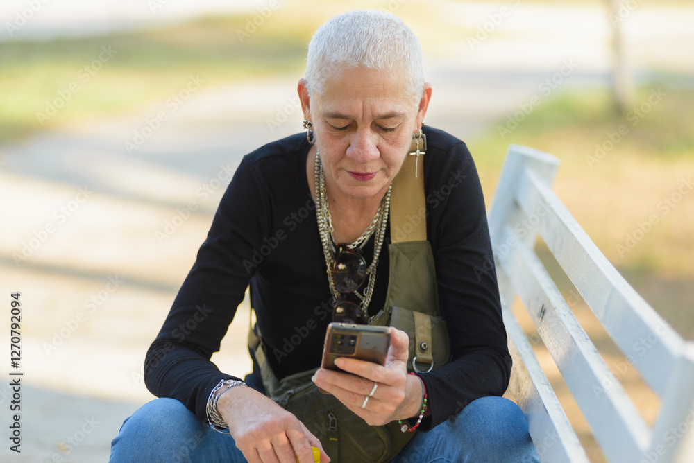 Fototapeta premium Middle-aged lesbian woman sitting on a park bench checks her cell phone.