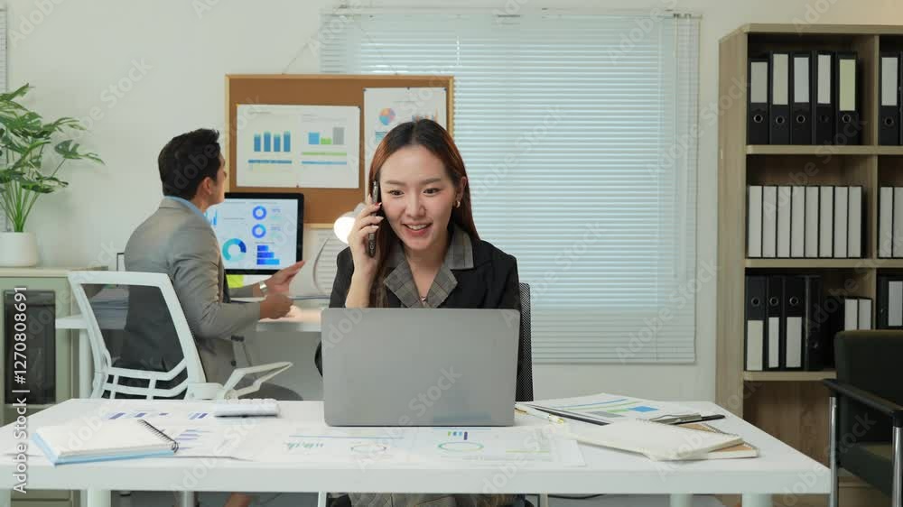 Professional businesswoman communicating via phone while working on laptop, colleague collaborating in background within modern office environment Professional businesswoman communicating via phone while working on laptop, colleague collaborating in background within modern office environment