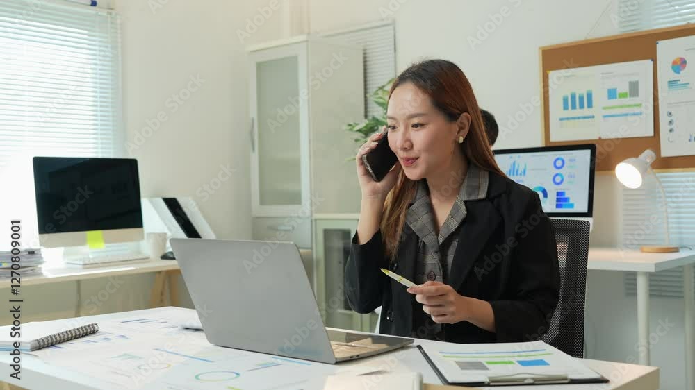 Professional businesswoman analyzing financial charts while talking on phone, male colleague working on laptop in modern, bright office workspace