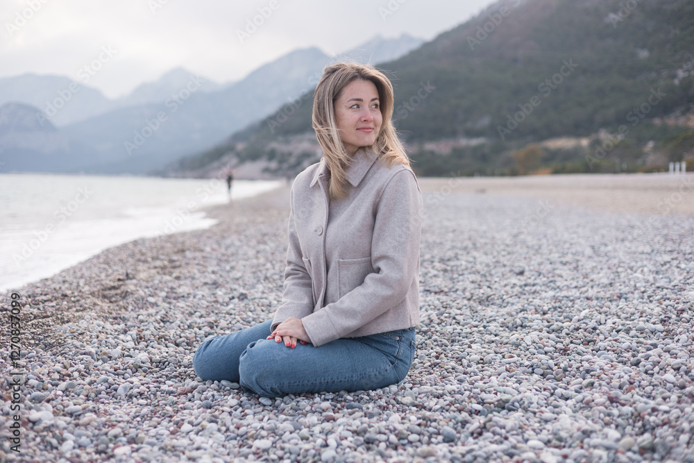 Blonde woman sits on the pebbled beach in winter Blonde woman sits on the pebbled beach in winter