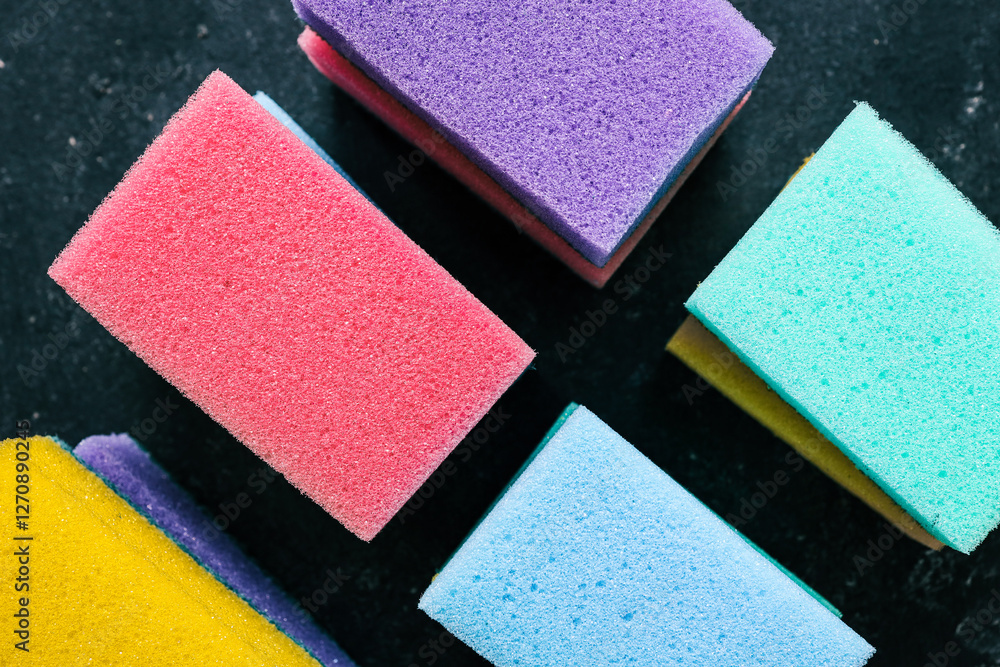 Fototapeta premium Colorful dishwashing sponges with scrubbing layers arranged in a row on a dark textured background. Household cleaning tools used for washing dishes, scrubbing surfaces, and removing dirt