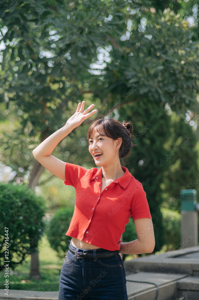 Asian cheerful woman in red shirt and jeans waves her hand while standing outdoors in sunny park surrounded by greenery Asian cheerful woman in red shirt and jeans waves her hand while standing outdoors in sunny park surrounded by greenery
