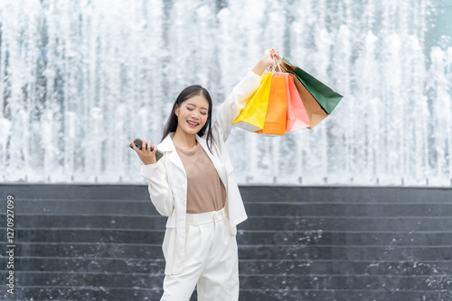 Fotografija Joyful Shopping Spree: A radiant woman beams with delight, hoisting vibrant shopping bags aloft, smartphone in hand, against the backdrop of an enchanting fountain