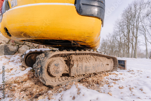 A close-up of a yellow excavator’s muddy tracks on a snowy construction site, showing dirt-covered treads and rugged terrain with a winter landscape in the background.. photo