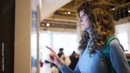Young woman with curly hair and backpack interacting with large touchscreen display in contemporary public space, exploring information with intuitive gestures