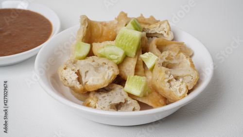 A portion of batagor (fried tofu meatballs) topped with sliced ​​cucumber served in a white container with peanut sauce in a separate container placed on a white base and background (isolated white). photo