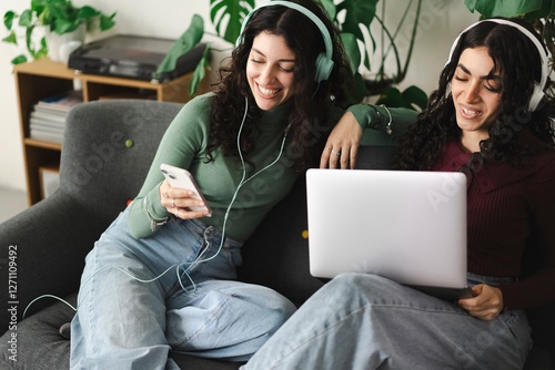 Two young women with curly hair are sitting on a sofa, one using a laptop while the other looks at her smartphone, both wearing headphones and enjoying a relaxing moment together. photo