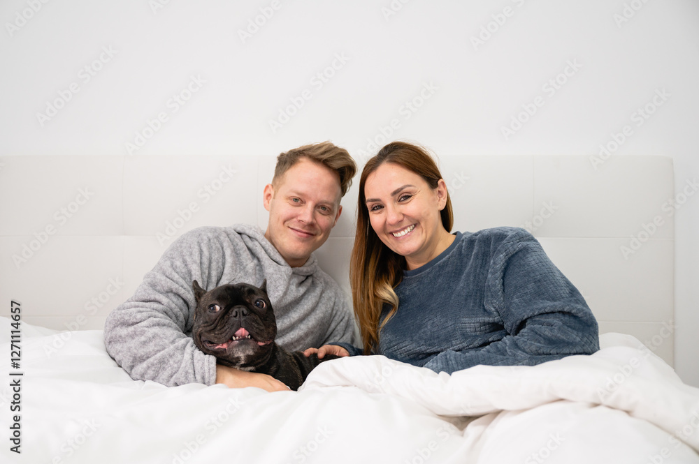 Loving couple are looking at the camera relaxing on the bed with their French bulldog with copy space Loving couple are looking at the camera relaxing on the bed with their French bulldog with copy space