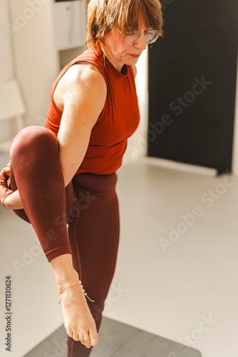 A woman in a red top and leggings performs a standing yoga pose, balancing on one leg while holding her foot, with a focused expression in a bright indoor space. photo