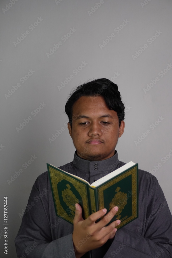 Southeast Asian Muslim man wearing grey robe holding and reading Quran carefully Southeast Asian Muslim man wearing grey robe holding and reading Quran carefully