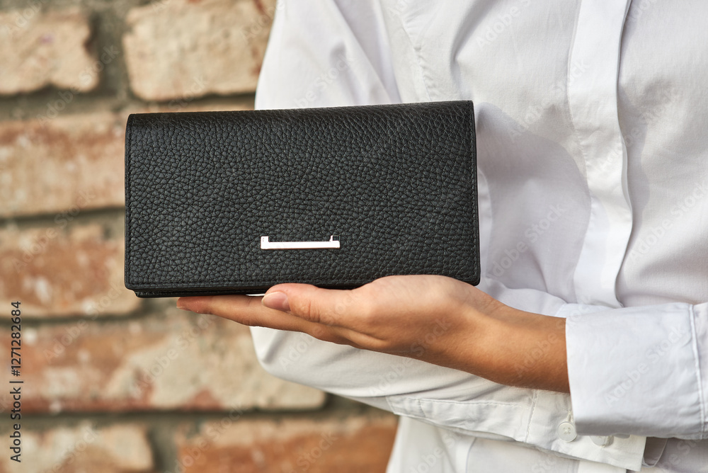 Elegant black leather wallet held by person in white shirt against brick wall, showcasing style and sophistication concept