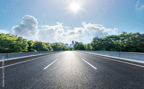 An asphalt road leads to the city through the forest and sunny sky
