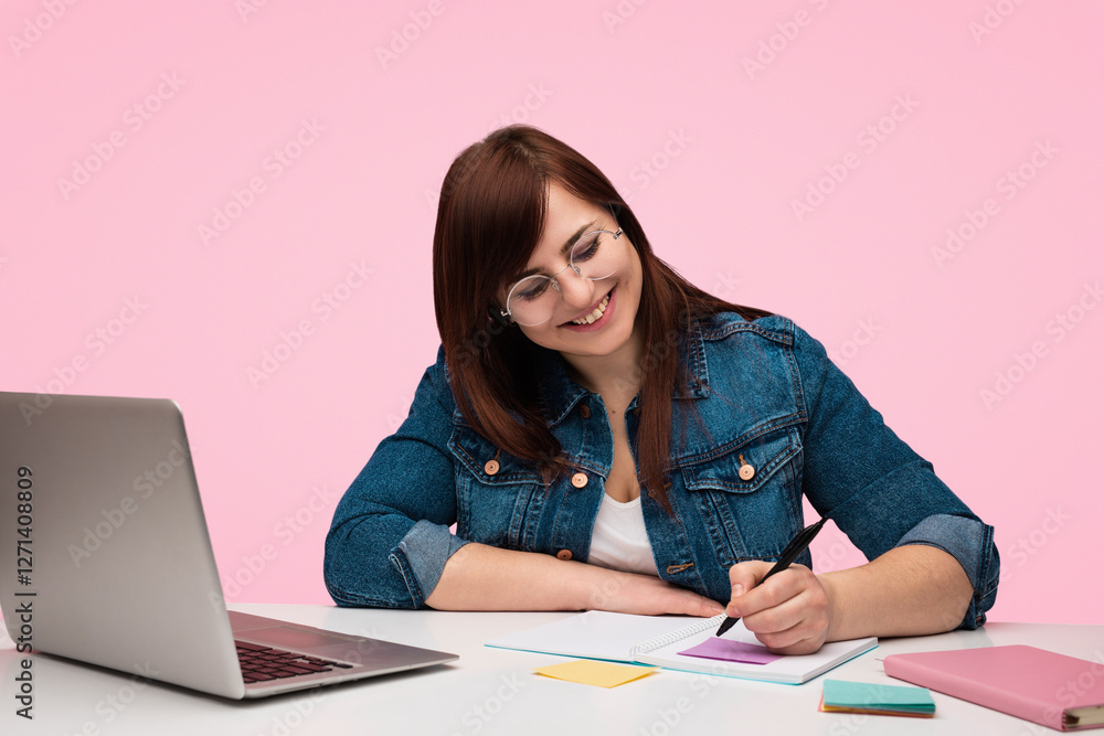 Confident woman in casual attire with glasses writing notes near a laptop, expressing focus and creativity against a pink background. Ideal for concepts of productivity, positivity, and personal