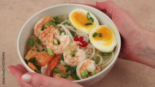 Asian dish with broth, noodles, tiger shrimp, fish, egg and green onions. The woman places the bowl on the table. Traditional Asian soup in a white deep plate. Selective focus, close-up.