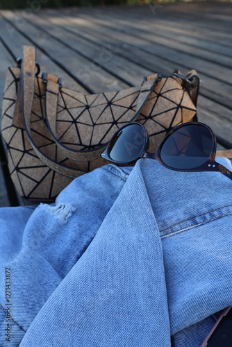 Sunglasses, denim jacket and women's handbag on wooden surface outdoors. Women's accessories