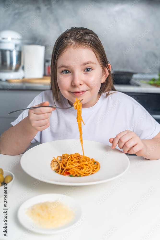 Cute little teen girl eating spaghetti on the background of gray kitchen at home sitting in white t-shirt at table.