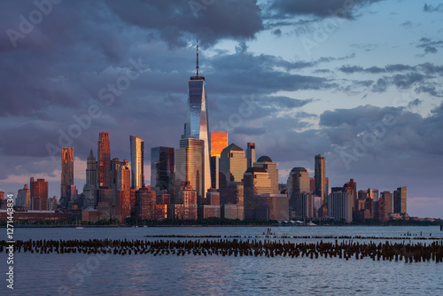 New York City skyline at sunset with Lower Manhattan and World Trade Center skyscrapers illuminated by golden light. Urban landscape across the Hudson River with Financial District and Battery Park