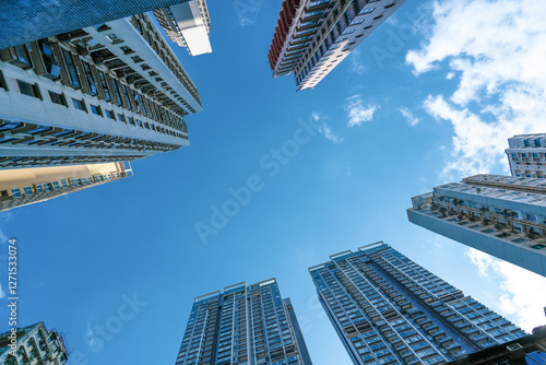 Tall skyscrapers line the streets of a bustling city, their glass facades reflecting the sunlight and bright sky above. Fluffy clouds drift peacefully overhead