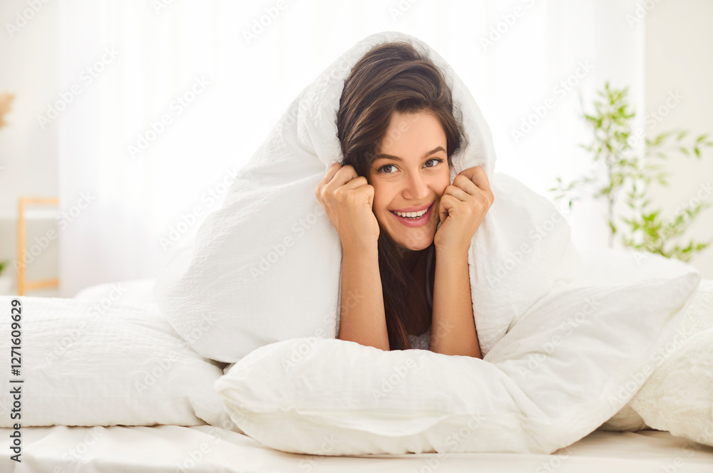 Pretty young woman lying in cosy comfortable bed, covering her head with white blanket. Smiling brunette girl relaxing in bedroom in early morning, enjoying leisure at home. Good morning concept. Pretty young woman lying in cosy comfortable bed, covering her head with white blanket. Smiling brunette girl relaxing in bedroom in early morning, enjoying leisure at home. Good morning concept.