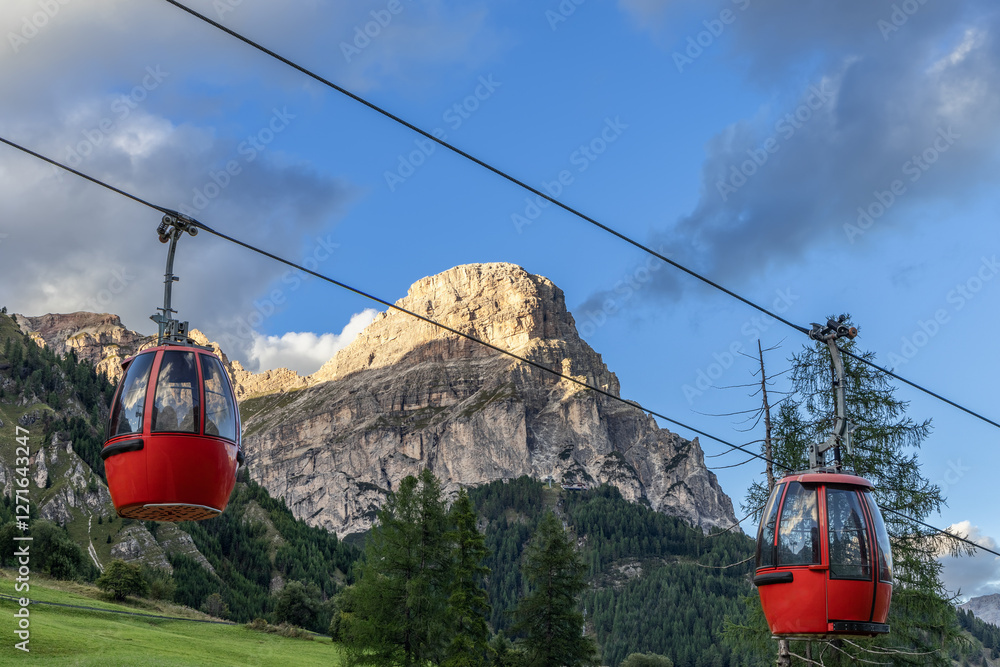 Red gondolas on a cableway system traverse the Dolomites in Italy, framed by the majestic Sassongher peak and vibrant greenery under soft evening light. Red gondolas on a cableway system traverse the Dolomites in Italy, framed by the majestic Sassongher peak and vibrant greenery under soft evening light.