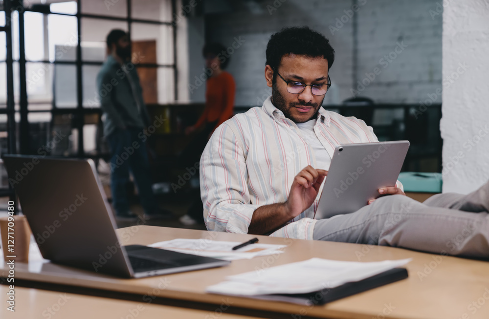 © BullRun - Focused ethnic man working on tablet while sitting at table with laptop © BullRun - Focused ethnic man working on tablet while sitting at table with laptop