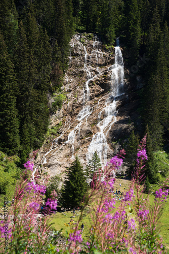 Scenic view of Brochaux waterfall  through blurry wild flowers in French Alps in summer near Avoriaz in Morzine area, Haute-Savoie, Auvergne-Rhone-Alpes, France. Silhouettes of relaxing tourists