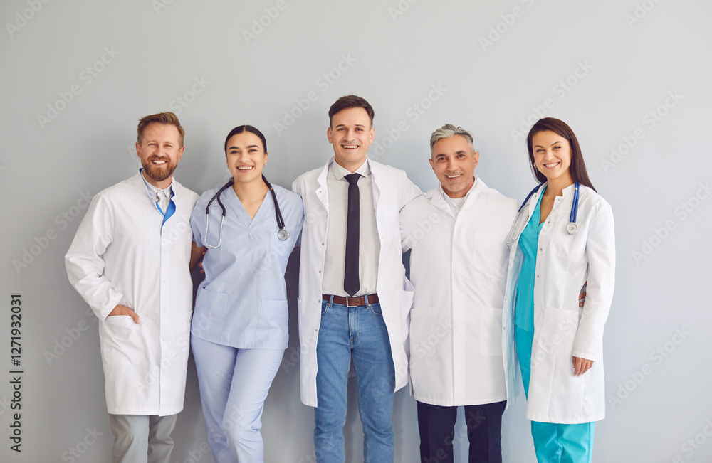 Group of young and adult doctors clinicians and cardiologists in scrubs and white coats standing together, hugging each other and smiling at camera on gray background. Happy clinic or hospital staff. Group of young and adult doctors clinicians and cardiologists in scrubs and white coats standing together, hugging each other and smiling at camera on gray background. Happy clinic or hospital staff.