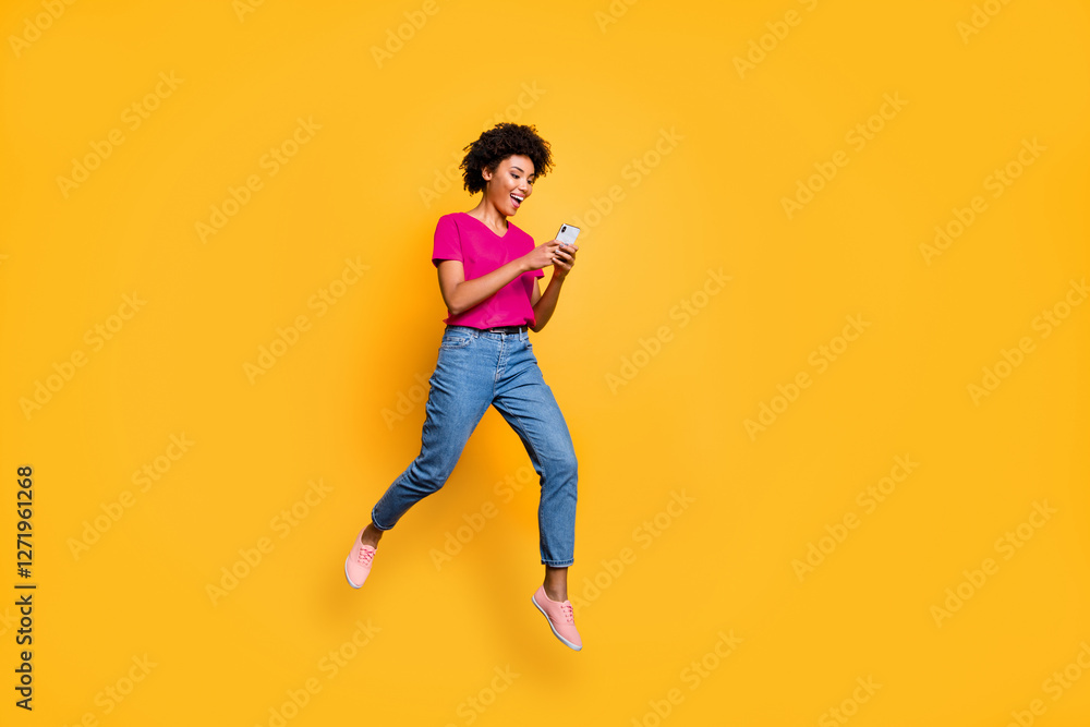 Full length photo of positive cheerful funny curly brown hair girl use smartphone read post blogs in social network jump wear red t-shirt denim jeans isolated over yellow color background Full length photo of positive cheerful funny curly brown hair girl use smartphone read post blogs in social network jump wear red t-shirt denim jeans isolated over yellow color background