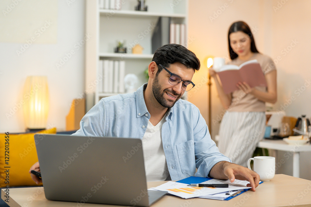 Young businessman working at home office on laptop computer, finishing his online work, while his girlfriend and roommate sitting behind on sofa relaxing and waiting. Freelance business. couple. Young businessman working at home office on laptop computer, finishing his online work, while his girlfriend and roommate sitting behind on sofa relaxing and waiting. Freelance business. couple.