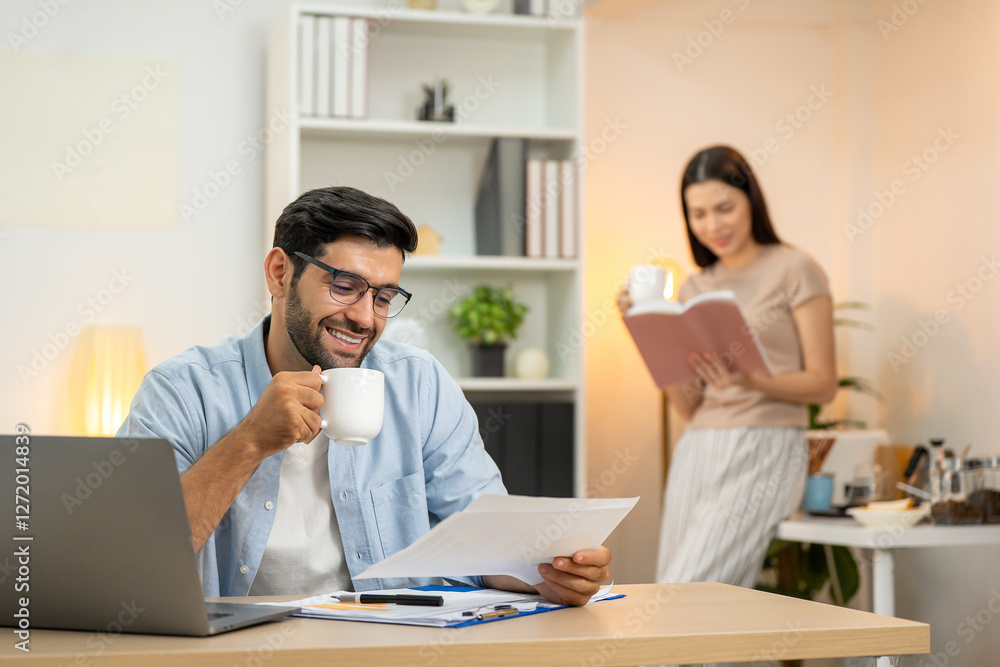 Young businessman working at home office on laptop computer, finishing his online work, while his girlfriend and roommate sitting behind on sofa relaxing and waiting. Freelance business. couple. Young businessman working at home office on laptop computer, finishing his online work, while his girlfriend and roommate sitting behind on sofa relaxing and waiting. Freelance business. couple.