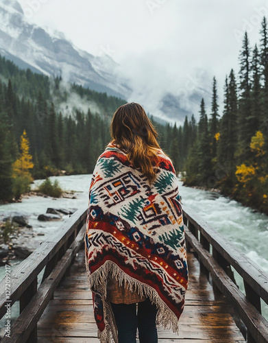 A woman wrapped in a pure white blanket with colorful black and red Native American patterns stands on a wooden bridge over a river against a backdrop of high mountains.