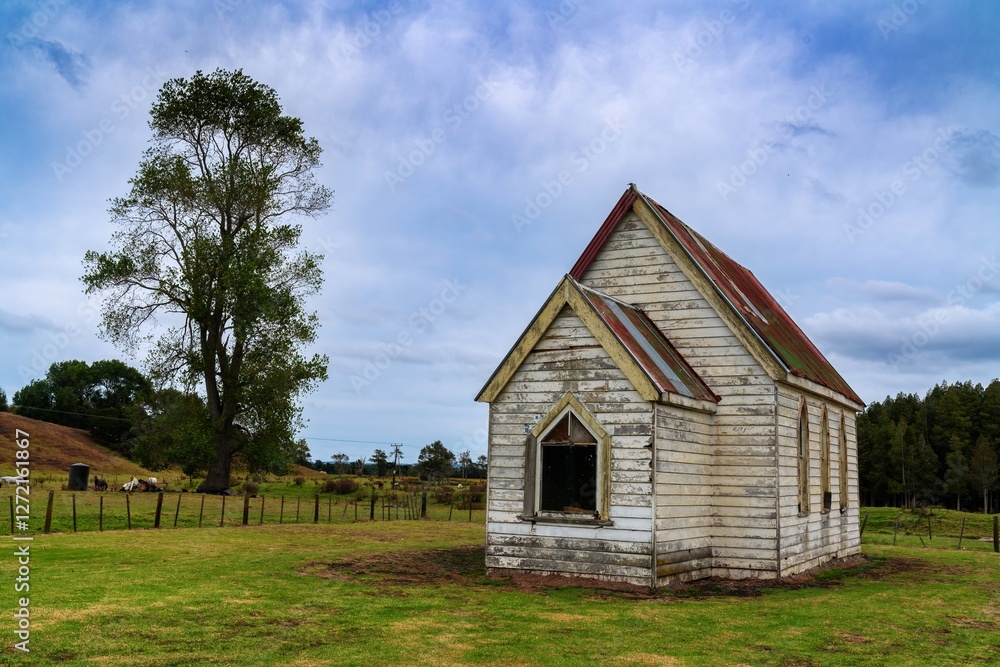 Obraz premium Abandoned church on a rural property. Horses graze nearby. A weathered, white wooden structure, with a faded red metal roof. Ruawai, Dargaville, Northland, NZ