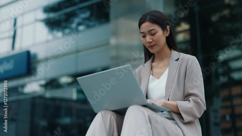 Portrait of Thai businesswoman working remotely on laptop sitting outside near office in downtown.